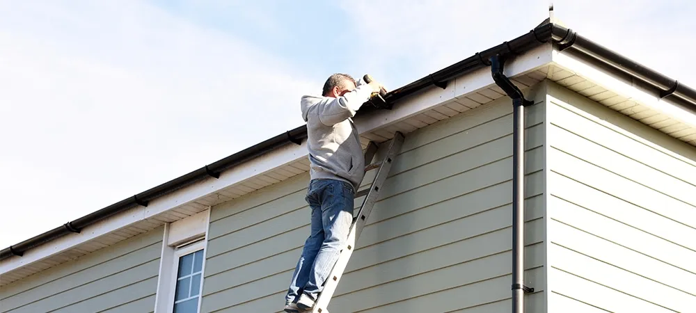 man on ladder cleaning house gutters during home maintenance or roof inspection task