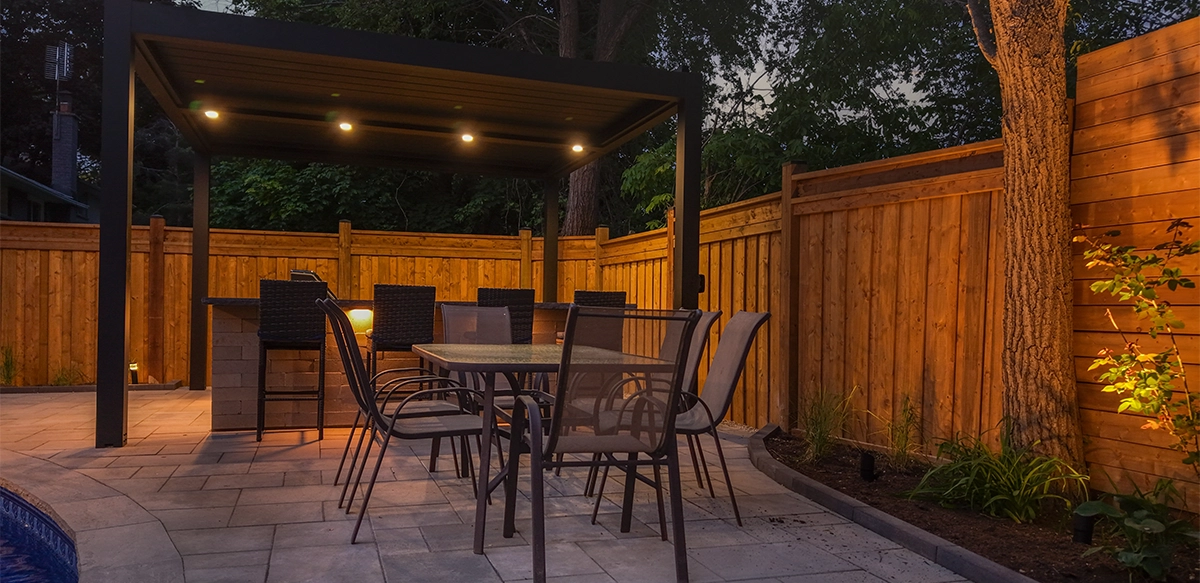 backyard lighting on at night, open kitchen with patio cover and dinner area. interlocking floor and fence with landscaping