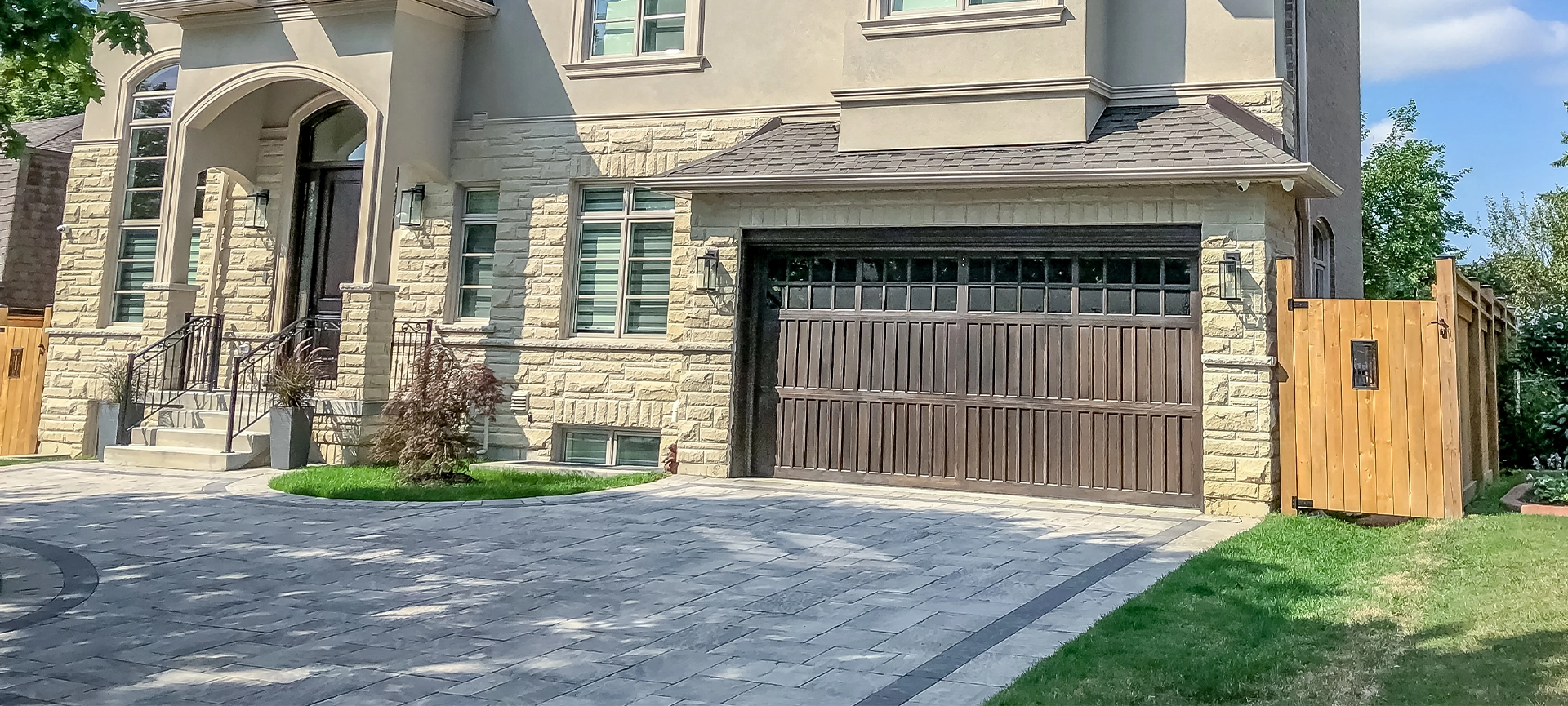 A beautiful large modern custom executive residential house with grey stone and brick, with gray shingles and grey trim, patterned interlock walkway and driveway, and double garage doors.