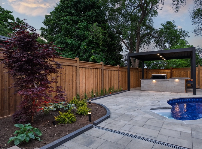home backyard with surrounding fence, open kitchen, landscaping and interlocking patio around pool