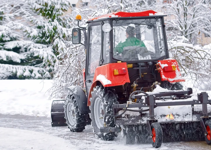 Tractor clear snow in park, machinery cleaning equipment. Snow removal red tractor clean walkway with plough sweeping brush. Tractor removing snow, cleaning sidewalk from snow in snowy winter