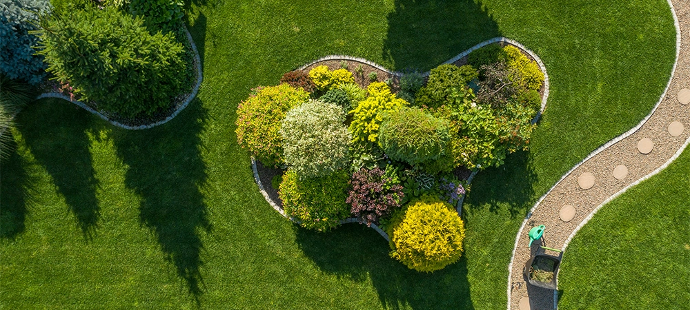 Garden Maintenance and Care Work in Process. Wheelbarrow with Grass Cuttings and a Plants Watering Can Stand on a Twisting Garden Path Next to a Large Landscape Island. Aerial View.