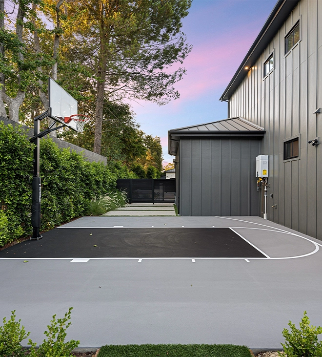 well maintained sports court A modern residential backyard with a basketball court, surrounded by lush greenery and a contemporary house.