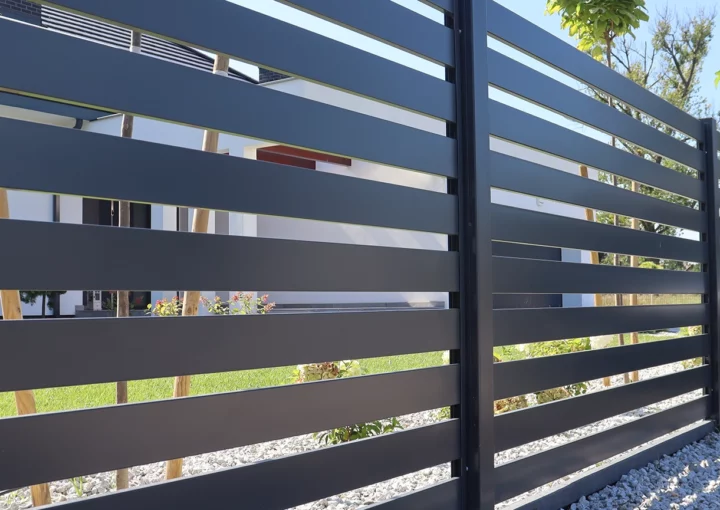 A sleek horizontal slat fence in a dark gray finish lines the perimeter of a modern home, complemented by decorative gravel and neatly planted ornamental trees under a clear blue sky.
