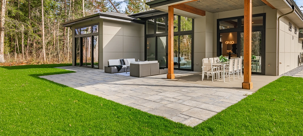 Exterior image of a contemporary home with flat roof and brown trim and lush green grass blue sky forest in the background furnished patio for outdoor dining and lounging