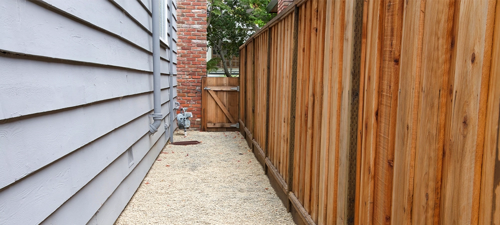 Long narrow side yard, new wood fence on the right with house on the left, home paint chipping and peeling. Meter by brick chimney exterior, fence to front yard with trees