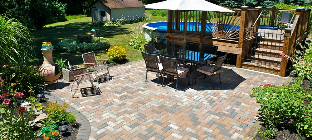 Beautiful backyard landscape featuring a brick paved patio with a dining set and umbrellas next to an above ground swimming pool with a wooden deck.