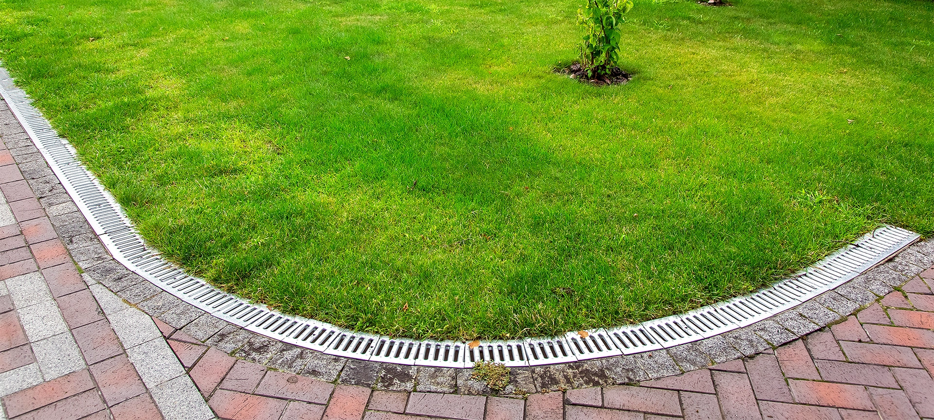 curved iron grid drainage system in the backyard by the green lawn with tree and footpath of red paving slabs.
