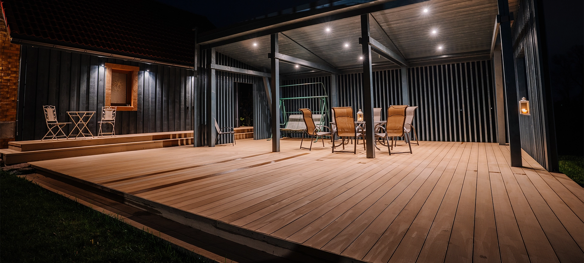 A modern wooden terrace at night with outdoor seating, a pergola, and warm lighting. A cozy ambiance with a covered dining area, a swing, and decorative lanterns.
