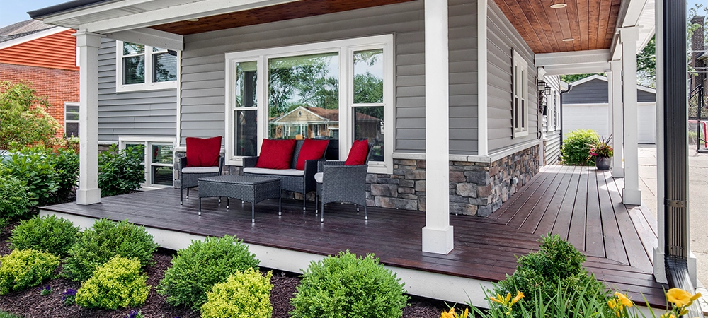 The exterior of a grey modern, suburban home with a covered porch and furniture featuring red accent pillows.